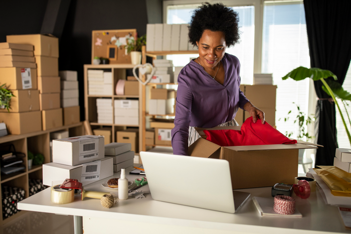 Businesswoman using laptop while packing clothes in a cardboard box