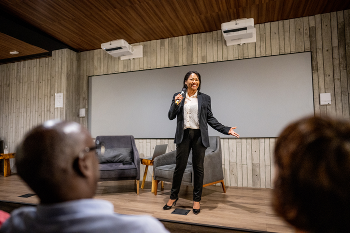 A small group of business professionals sit around a boardroom table as they meet to discuss new strategies.  They are each dressed professionally and are taking notes on laptops and in notebooks as a female manager leads the meeting.