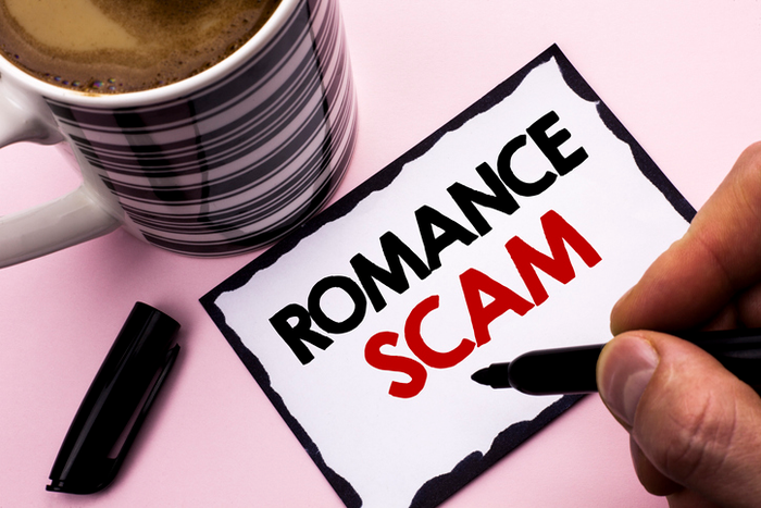 Person writing "Romance Scam" on piece of paper next to a mug of coffee