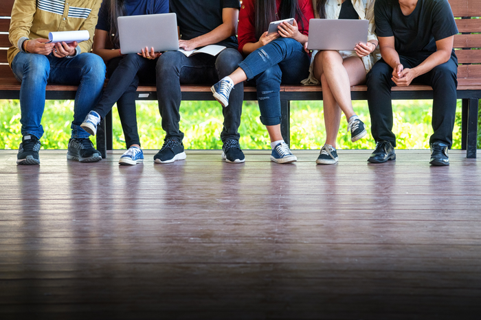 shows row of students sitting on a bench... just feet and legs and hands. some with computers or notepads.