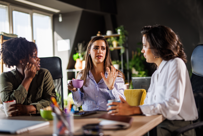 Three diverse young women sitting at table, enjoying coffee and chatting