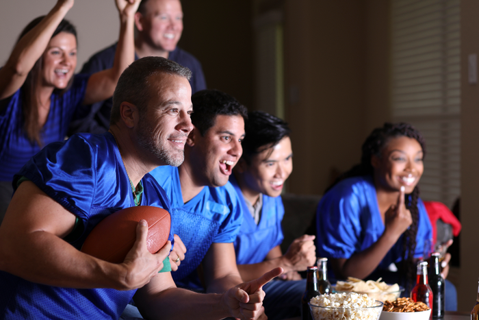 group of football fans watching the game together on television at home.  They have snacks, drinks, and sports ball and wear blue team jerseys.  