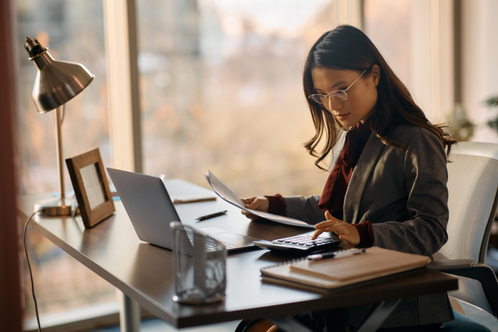 Asian female entrepreneur using calculator while going through business reports in the office.