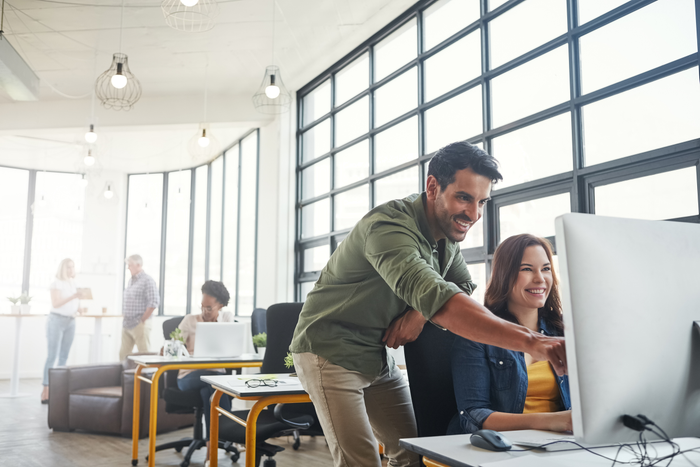 Man and woman work together at a computer with coworkers in the background.