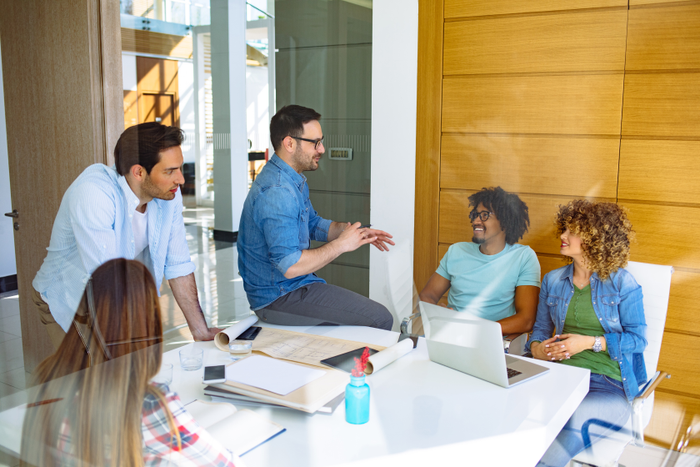 A small group of business professionals sit around a boardroom table as they meet to discuss new strategies.  They are each dressed professionally and are taking notes on laptops and in notebooks as a female manager leads the meeting.