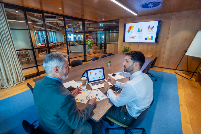 Two business men meeting in a board room. There is technology and documents on the conference table including a laptop