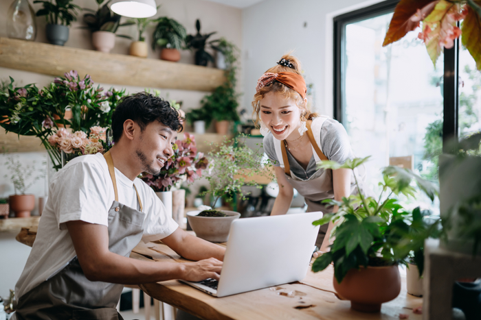 Smiling young Asian couple, the owners of small business flower shop, discussing over laptop on counter against flowers and plants. Start-up business, business partnership and teamwork. Working together for successful business