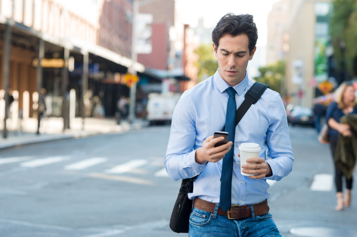 Businessman using smartphone and holding paper cup ina urban scene. Worried businessman in walking on the road and messaging with phone. Young man text messaging through cell phone while walking on the road in the city centre.