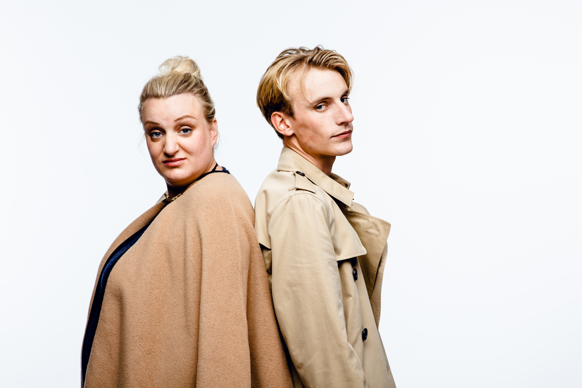 A blonde woman with hair tied in a bun and wearing a camel coloured cape, standing back to back with a blond man wearing a beige trench coat.