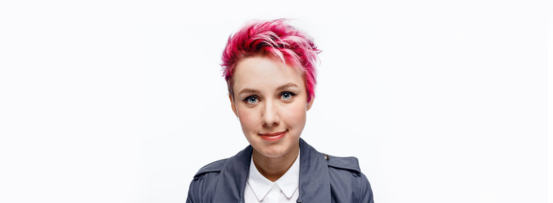 A woman with a dyed pink pixie crop wearing a white shirt and grey trench coat.